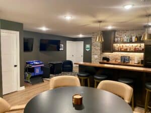 A spacious finished basement featuring a dark wood home bar with a brick backsplash and illuminated liquor shelves. The room is designed for entertainment, including a round table with tan chairs in the foreground, two wall-mounted TVs, and a "Golden Tee" arcade game near several white interior doors.