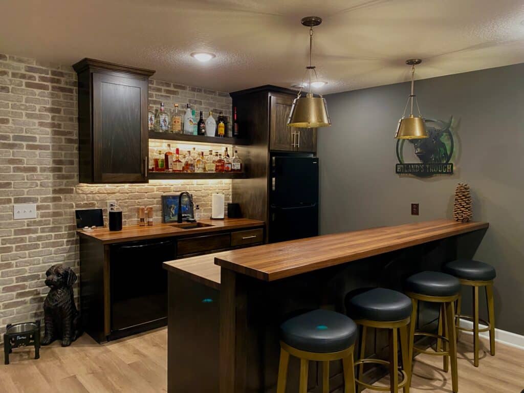 Updated rustic home bar featuring a brick accent wall, dark wood cabinetry, and a butcher block bar top with four leather-seated stools. The space includes illuminated floating shelves displaying various liquor bottles, brass pendant lights, and a small black refrigerator.