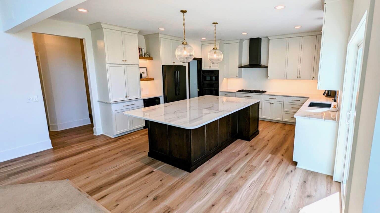 A wide-angle view of the kitchen featuring a dark wood island with a white marble countertop and two glass globe pendant lights. The space includes white perimeter cabinetry, a black range hood, stainless steel appliances, and light-toned hardwood flooring throughout.